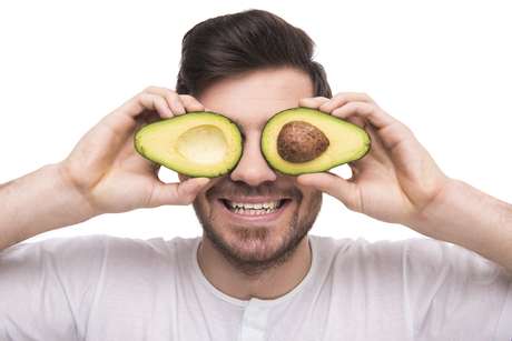 Young man is holding avocado in front of his eyes isolated on white background.